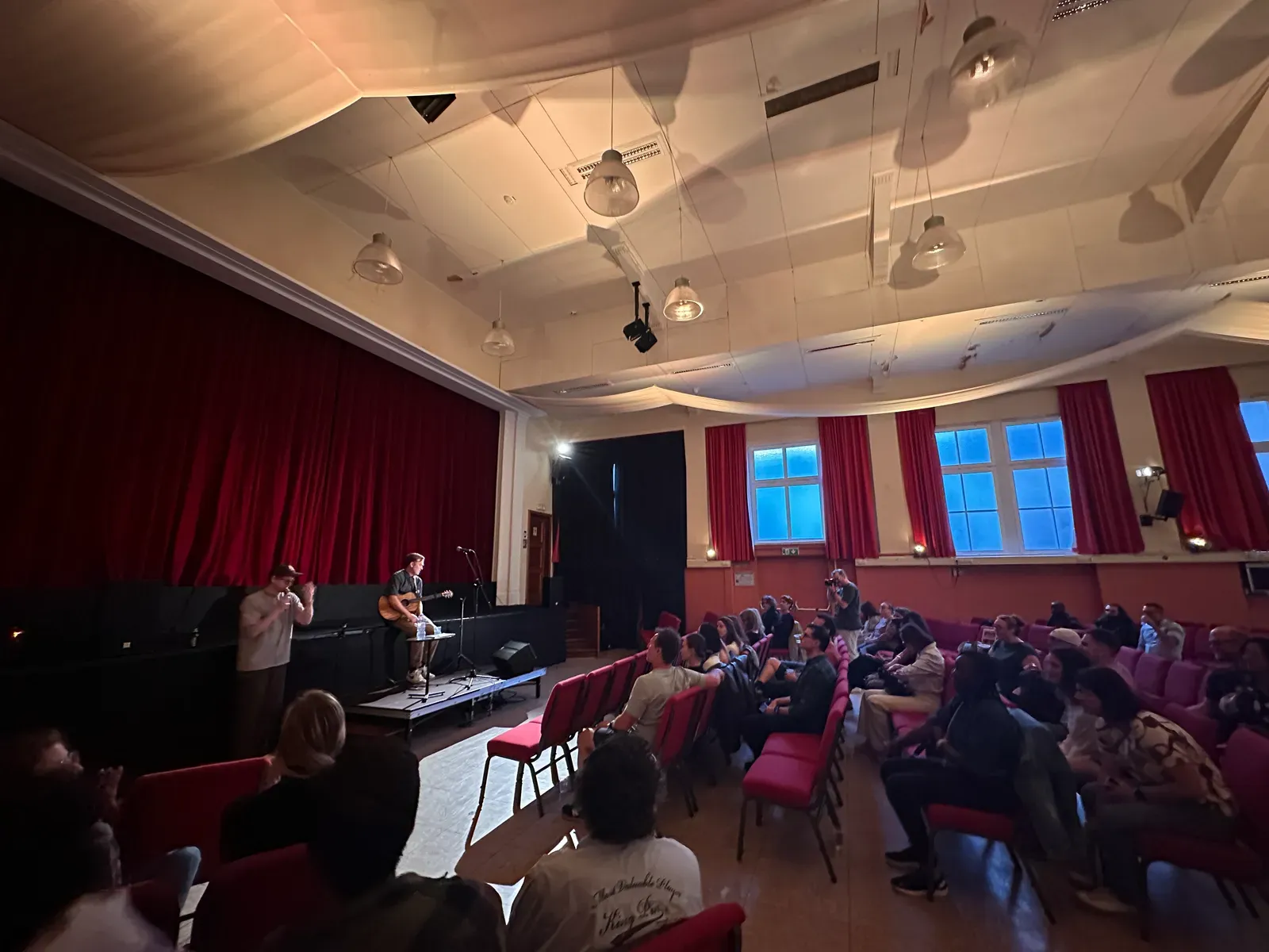 Der SSJ-Saal voller Gäste mit Blick zur Bühne, rote Stühle und Vorhänge, Andy an der Gitarre